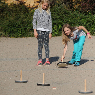 Deux enfants jouent au curling sur un terrain en gravier. Une fille aux cheveux longs et blonds se penche pour lancer un disque avec une poignée, tandis que l'autre fille, également blonde et portant un vêtement à motifs léopard, observe. Des disques sont