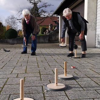 Deux femmes âgées jouent au curling sur un sol en pavés, chacune tenant un équipement de jeu. Elles sont concentrées sur le lancer des pièces de jeu, avec plusieurs cibles en bois en vue sur le sol. L'environnement est extérieur, avec des bâtiments et des