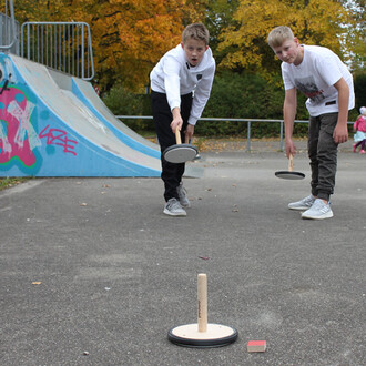 Deux jeunes garçons jouent au curling avec des disques en bois sur une surface en béton. L'un des garçons est en position de lancer son disque, tandis que l'autre attend son tour. En arrière-plan, on aperçoit un skatepark et des arbres aux feuillages colo