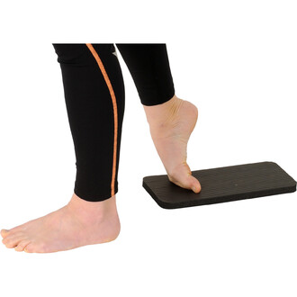 Pied nu posé sur une planche noire, avec l'autre pied à proximité, montrant un exercice d'équilibre. La personne porte un pantalon noir avec une décoration orange sur le côté.