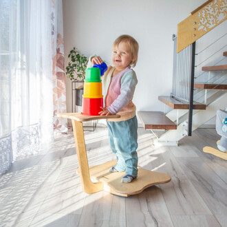 Un enfant debout sur une planche en bois, souriant tout en empilant des gobelets colorés (rouge, jaune, vert et bleu) sur la planche. En arrière-plan, on voit des rideaux légers et une plante verte. L'intérieur est bien éclairé par la lumière naturelle.