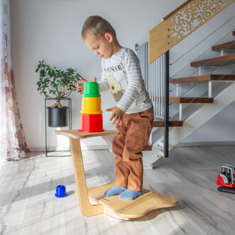 Un jeune enfant se tient debout sur une plateforme en bois, en train d'empiler des tasses colorées (vert, jaune, rouge) en plastique. À l'arrière-plan, on aperçoit une plante dans un pot noir et un escalier. Le sol est en bois clair, et un petit jouet est