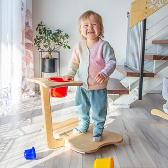 Enfant joyeux debout sur un plateau en bois, souriant largement, portant un pull coloré. Il manipule un seau rouge, avec des blocs de couleurs dispersés autour de lui dans une pièce lumineuse. Une plante est visible en arrière-plan.