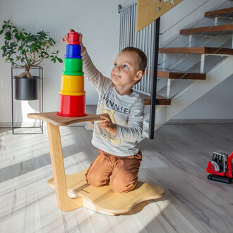 Un enfant est à genoux sur un support en bois, en train d'empiler des gobelets colorés. Il tient le gobelet rouge en haut de la pile, qui se compose également de gobelets bleus, verts, jaunes et rouges. En arrière-plan, on voit une plante en pot et un esc