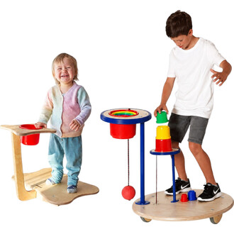 Deux enfants jouent avec le Pedalo Fun-Wipp : une fillette sur une planche en bois avec un bol rouge et un garçon près d’une structure bleue à plateau arc-en-ciel et pièces colorées (rouge, jaune, bleu) sur socle en bois.