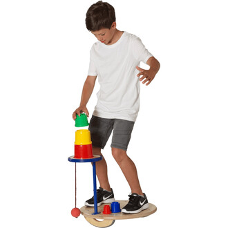 Un enfant se tient debout sur une planche d'équilibre en train de superposer des gobelets colorés. Il porte un t-shirt blanc et un short gris, et il semble concentré sur la tâche. Les gobelets empilés incluent des couleurs vertes, jaunes et rouges. Au bas