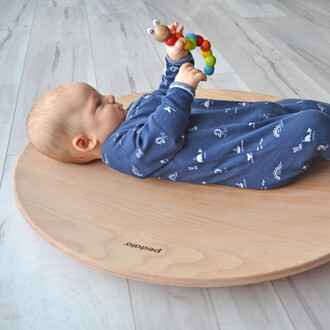 Un bébé allongé sur un plateau en bois, tenant un jouet coloré en forme de chenille. Le bébé porte un pyjama bleu à motifs et regarde le jouet avec intérêt. Le fond est composé de parquets clairs.