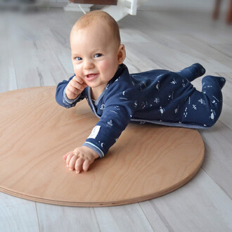 Bébé souriant en tenue de nuit bleu marine, allongé sur un tapis en bois rond, avec un bras tendu et l'autre main sur le menton, exprimant de la curiosité et de la joie.