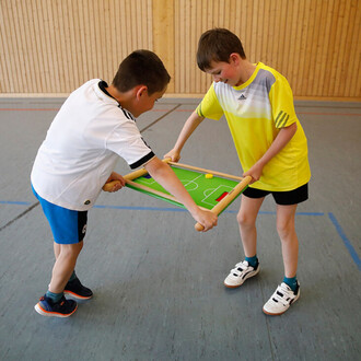 Deux enfants jouent ensemble à un jeu de football sur une table, chacun tenant un côté d'un plateau de jeu. L'un d'eux porte un maillot blanc, tandis que l'autre est vêtu d'un T-shirt jaune. Le plateau de jeu est vert avec un terrain de football dessiné, 