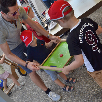 Des enfants jouent à un jeu de table représentant un terrain de football, avec un adulte les aidant à tenir le plateau. Les enfants portent des casquettes rouges, l'un est en t-shirt noir, l'autre en maillot de football. Le plateau est vert avec un petit 