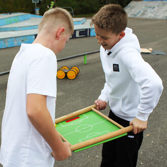 Deux garçons jouent avec un petit terrain de football en bois, souriant et concentrés. Le terrain est vert avec des zones marquées, et ils tiennent le cadre en bois chacun d'un côté. En arrière-plan, on aperçoit un skatepark et des accessoires de jongleri