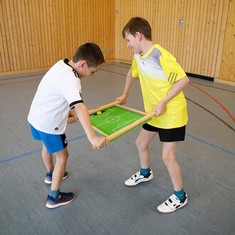 Deux enfants s'amusent dans une salle de sport en jouant à un jeu de football sur une table miniaturisée. Ils tiennent chacun un côté d'un cadre en bois, avec un terrain de football vert au milieu. L'un porte un maillot blanc et l'autre un maillot jaune. 