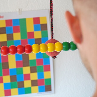 Vue rapprochée d'un accessoire de jonglerie, montrant des perles colorées (rouges, jaunes, vertes) fixées sur une corde, avec un fond de mural décoré de carrés colorés (bleu, vert, jaune, rouge et marron). Une partie d'une oreille est visible à droite de 