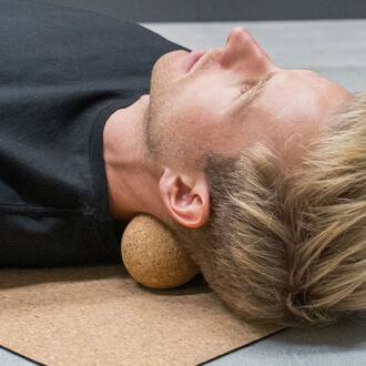 Homme allongé sur le dos, la tête reposant sur une boule de massage en liège, sur un tapis en liège, avec une expression de détente.