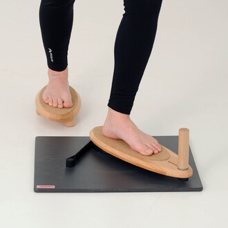 Pieds placés sur des équipements d'équilibre en bois, incluant une plateforme avec une base noire, sur laquelle repose un accessoire de jonglerie. Un pied est sur un support arrondi, tandis que l'autre est sur une planche légèrement inclinée. Les jambes p