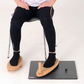 Image d'un pratiquant assis sur une chaise avec des jambes en leggings noirs. Ses pieds sont placés sur deux supports en bois avec une base noire. Les supports ont une surface en liège et un élément vertical en bois. Le décor est neutre, mettant en valeur