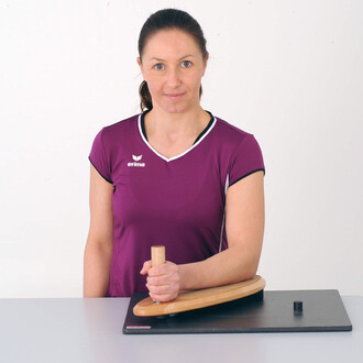 Femme en t-shirt violet tenant un dispositif de jonglerie sur une table, avec un fond blanc. Le dispositif est en bois avec une partie courbée et un petit cylindre noir à côté.