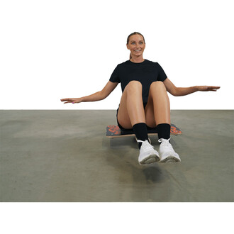 Femme assise sur une planche de jonglerie, les bras tendus sur les côtés, souriant. Elle porte un t-shirt noir, un short, et des chaussettes noires, avec des chaussures blanches. Le sol est en béton.