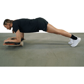 Femme en position de planche sur un matériel de fitness, avec les avant-bras posés sur la planche et le corps aligné. Elle porte un vêtement de sport noir et des chaussures blanches, sur un sol gris.