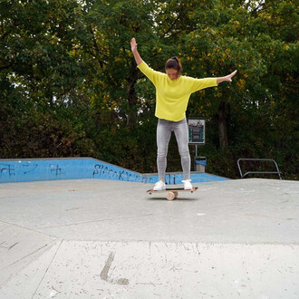 Femme en pull jaune, en train d'équilibrer sur une planche de skate, dans un skatepark, les bras levés pour maintenir son équilibre, entourée d'arbres verdoyants.