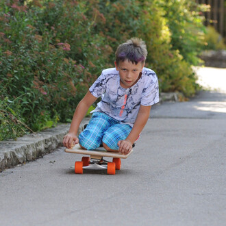 Un garçon en train de glisser sur un skateboard, positionné à plat sur la route. Il porte un t-shirt à motifs et un short à carreaux. Son expression est concentrée et déterminée, tandis qu'il avance sur une surface pavée entourée de verdure.