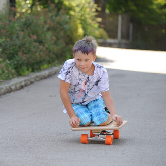 Un enfant s'amuse en position assise sur une planche à roulettes, sur une route ensoleillée bordée de verdure. Il porte un t-shirt à motifs marbrés et un short à carreaux. Les roues de la planche sont orange.