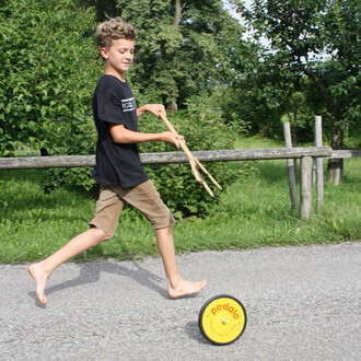 Un enfant en t-shirt noir court pieds nus sur une route, tenant des bâtons. Il s'amuse avec un jouet roulant de couleur jaune portant le logo pedalo. En arrière-plan, on distingue une verdure dense et une barrière en bois.