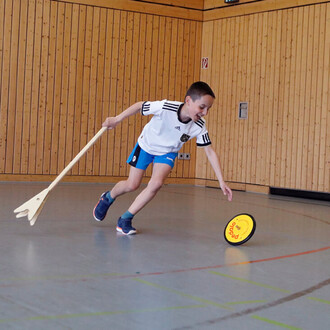 Un enfant courbe le dos, vêtu d'un maillot blanc avec des rayures noires et d'un short bleu, en train d'attraper un disque jaune sur le sol à l'aide d'une grande pagaie en bois. L'image se déroule dans une salle au sol en sport, avec des murs en bois clai