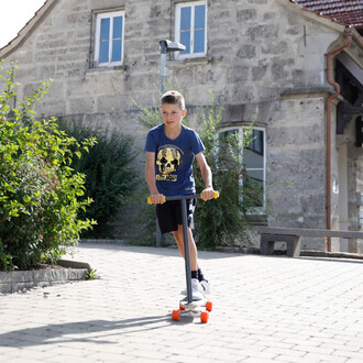 Enfant sur trottinette, portant un t-shirt bleu avec un motif de skull, roulant sur un chemin pavé, entouré de verdure et d'une maison en arrière-plan.