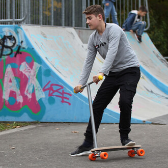 Un jeune homme roule sur un scooter à la surface d'un skatepark. Il porte un t-shirt gris et un pantalon noir. Le scooter a un plateau en bois et des roues oranges. En arrière-plan, on peut voir des rampes de skate avec des graffitis colorés. D'autres enf