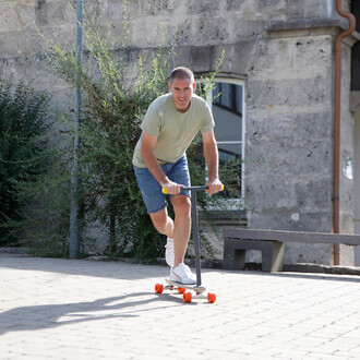 Homme souriant en train de rouler sur une trottinette, portant un t-shirt gris et un short en jean. Il se trouve sur un sol pavé, avec des plantes en arrière-plan et un bâtiment en pierre. La trottinette a des roues orange et un guidon noir.