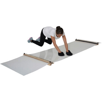 Femme en train de manipuler un tapis de jonglerie, à genoux, utilisant des gants noirs pour ajuster la surface. Le tapis est fixé entre deux barres en bois, posé sur un sol clair.