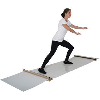 Femme debout sur un tapis de jonglerie, le corps en position dynamique, les bras tendus en avant. Elle porte un t-shirt blanc et un pantalon noir, avec des chaussures noires. Le tapis est transparent avec des supports en bois aux extrémités.