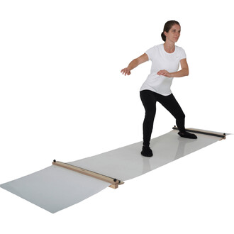 Femme en tenue de sport blanche, positionnée sur un tapis de jonglerie. Elle est inclinée légèrement en avant, les bras écartés pour l'équilibre, avec un regard concentré. Le tapis est en deux parties, fixées par des supports en bois, et se trouve sur un 