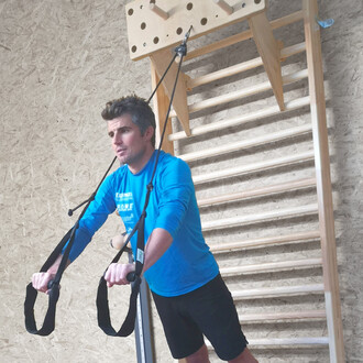 Un homme en vêtements de sport bleu effectue un exercice de traction à l'aide de sangles, debout devant un mur d'escalade en bois. Il semble concentré, avec des barres horizontales en arrière-plan.