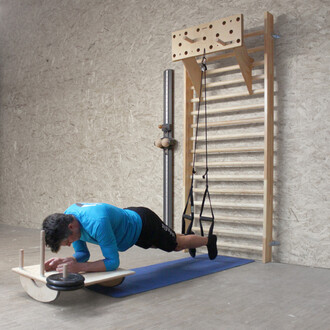 Un homme en position de planche est allongé sur un appareil de fitness en bois, avec des éléments de suspension attachés à un mur. Il porte un t-shirt bleu et un short noir. L'équipement est placé sur un tapis de sport, et des barres de gymnastique sont v