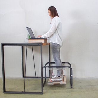 Femme debout devant un bureau, utilisant un ordinateur portable. Elle porte une chemise blanche et des jeans, et se tient sur une plateforme de soutien. Le bureau a un design moderne avec une structure en métal et une surface en bois, et une petite plante