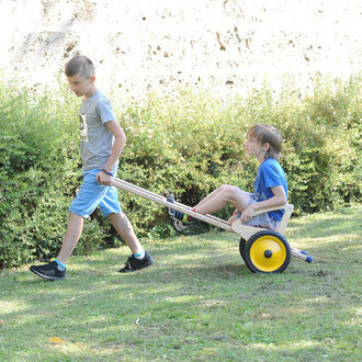 Deux enfants jouent dans un jardin. L'un tire un chariot en bois sur roues, tandis que l'autre est assis à l'intérieur. Le sol est enherbé et entouré de buissons verts. Les enfants portent des vêtements décontractés.