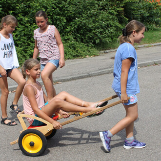 Quatre enfants jouent à l'extérieur. Une fille est assise dans un petit véhicule à roues en bois, tandis qu'une autre la tire en marchant. Deux autres enfants les regardent en riant. L'ambiance est enjouée et l'environnement est ensoleillé, avec des arbre