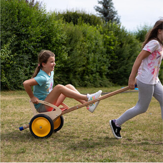 Deux enfants jouent à l'extérieur. L'un est assis dans une petite brouette en bois à refouler, riant joyeusement, tandis que l'autre tire la brouette par une barre. Le cadre est verdoyant, avec des buissons en arrière-plan. Les enfants portent des vêtemen