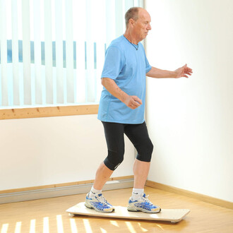 Homme âgé en t-shirt bleu et collants noirs, marchant sur une planche d'équilibre dans une pièce lumineuse. Des rideaux blancs en arrière-plan laissent passer la lumière.