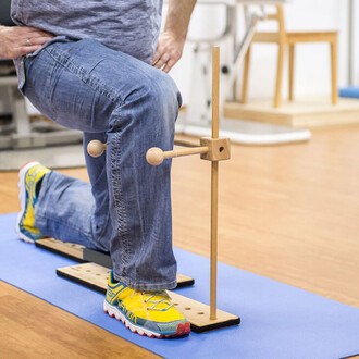 Un homme à genoux sur un tapis bleu, portant un t-shirt gris et un jean. Il utilise un accessoire de support en bois avec deux boules à chaque extrémité, positionné près de son genou. Ses chaussures sont multicolores, principalement jaunes avec des accent