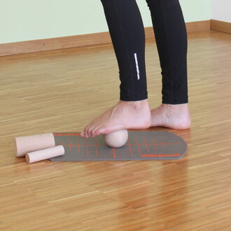 Une personne effectue un exercice sur un tapis de gymnastique. Elle est pieds nus, l'un de ses pieds repose sur une boule en bois, tandis que l'autre pied est légèrement levé. À côté de la boule, il y a deux cylindres en bois. Le sol est en bois clair et 