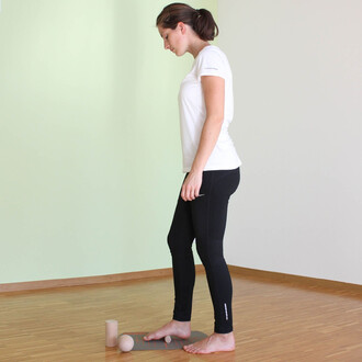 Femme debout sur un tapis, pieds nus, en t-shirt blanc et leggings noirs, regardant vers le sol. À ses pieds, des accessoires de jonglerie : une balle et un objet cylindrique. Le fond est de couleur claire, et le sol est en bois.