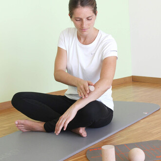 Femme assise sur un tapis, portant un t-shirt blanc et un pantalon noir, appliquant une technique de soin sur son bras avec un petit rouleau. En arrière-plan, un espace intérieur avec un mur vert pâle et un sol en bois, ainsi que des accessoires de jongle