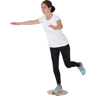 Femme en tenue de sport blanche et noire effectuant un exercice d'équilibre sur une plateforme ronde en bois, un bras tendu en avant. Elle porte des chaussures de sport bleu clair avec des éléments rose.