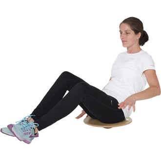 Femme assise sur un plateau d'équilibre en bois, portant un T-shirt blanc et un pantalon noir, avec des chaussures de sport bleu et violet. Elle utilise ses mains pour se stabiliser tout en s'amusant à l'exercice d'équilibre.