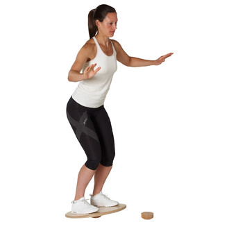 Femme en tenue de sport, se tenant en équilibre sur un plateau de balance en bois, les bras écartés pour maintenir sa stabilité. Elle porte un débardeur blanc et un legging noir avec des motifs, ainsi que des chaussures de sport blanches. Un accessoire de