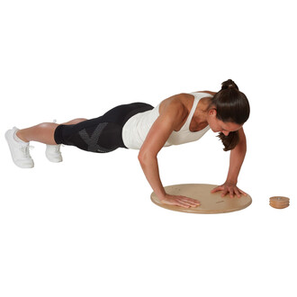 Femme en position de pompes sur un disque d'équilibre en bois, portant un débardeur blanc et un legging noir. Elle est concentrée, avec les mains posées sur le disque et le corps en ligne droite. Un accessoire rond en bois est visible à côté.