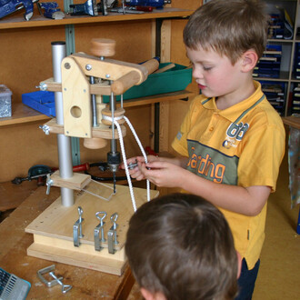 Un enfant utilisant un appareil de fabrication de jonglerie, manipulant des pièces métalliques et des cordes, avec un second enfant en arrière-plan. L'atelier est bien rangé, avec des outils et des matériaux visibles sur les étagères.